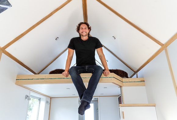 Photo of presenter Bryce Langston dressed in a black t-shirt and jeans, sitting on the floor of a loft in one of his own designs, a white and warm wood tiny house.
