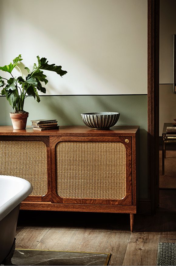 Photo of a sideboard made with cognac oak and rattan, dressed with a live plant and a bowl, in front of a grey-green painted wall with the top half of the wall an off-white. The sideboard sits atop brown wooden floorboards. 