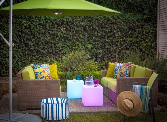 Photo of a small urban patio with a lime green umbrella overhead. Underneath, two tan textured outdoor loveseats with lime green seats and backs are dressed with multi-colored floral patterned throw pillows. A matching lime green and metal chair sits next to one loveseat, with a striped towel and sunhat hanging off one arm. A blue and white striped footstool sits opposite. Between the sofas are two LED-lit cubes, one in purple and one in blue, holding glasses and a jug of water. The backdrop for the entire space is a living wall of green vines.