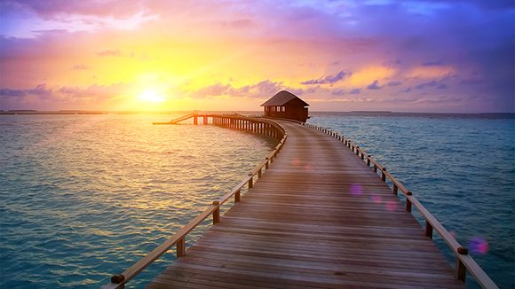 A beautiful sunset with pink, purple, yellow and orange, behind a single shack at the end of a long wooden dock over a blue ocean. Inspiration to quarantine staycation at home.