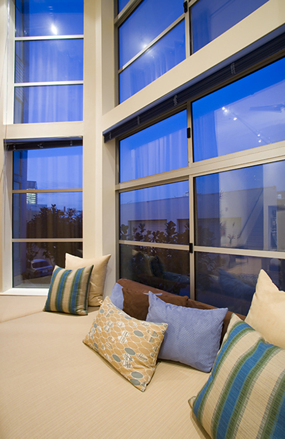 After the renovation, the full double-height windows are open and on view without window dressings, revealing a blue sky. Now the surface in front is a window seat of soft tan padded pillow, with multiple throw pillows in yellows, blues, and greens, some striped, some patterned, and some solid colors, stacked in front of the window.