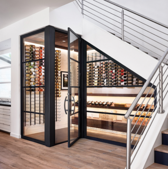 Under a white set of modern stairs with silver railings, is a black metal and glass enclosed storage area for wines, that also controls temperature and humidity with a glass door and LED lighting.