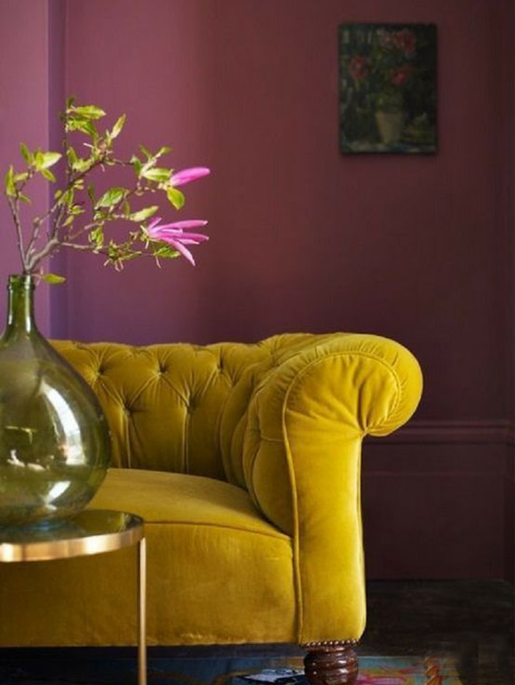 Photo of the end of a mustard yellow sofa with tufted back pillows and rounded arms on dark wooden round leg, in front of a mauve-pink wall color. The pink is echoed in a plant cutting in a large green glass vase on a brass and glass table in front.