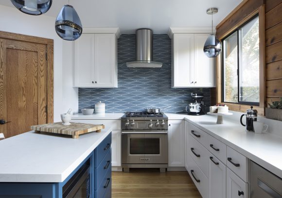 A contemporary kitchen with warm orangey wood doors and slatted wall around the sliding window. White countertops, drawers and cabinets have black handle pulls. Dark blue bottom cabinets on the kitchen island have matching handles. A silver oven and cooktop under a silver hood is surrounded by blue and white wavy backsplash tiles.