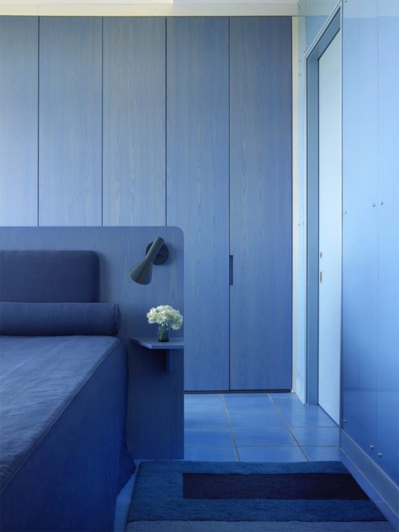 A bedroom designed in multiple shades and hues of blue, from floor tiles, to striped bedside rug, to wooden headboard with lamp, and blue pillows and sheets. A small white bunch of flowers sits on the bedside table nearly center of the image.