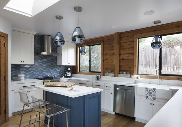 A beautiful contemporary kitchen in periwinkle blue and white, with knotty warm wood slatted walls. The upper and lower cabinets are white with dark metal drawer pulls and handles, and the countertops are white. The kitchen island has white countertop and periwinkle blue cabinetry. A matching blue and white patterned backsplash runs along the wall behind the cooktop and hood. A large white butler sink is in front of a picture frame window showing the backyard beyond.