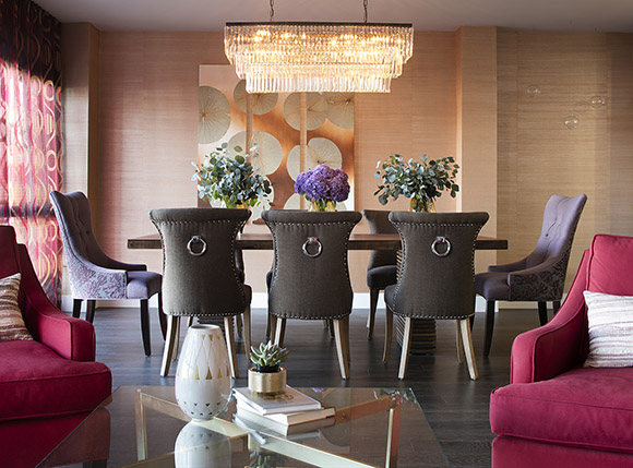 A contemporary dining room features a stunning rectangular glass tiered chandelier, over a dark wood rectangular table, surrounded by 6 modern dining chairs upholstered in brown fabric with nailheads and silver rings in the center of the backs. 2 head dining chairs are upholstered in a purple floral design on the backs and plain lilac tufted fabric on the inside. At the outer edges of the photo, two bright pink recliners are just visible, with a glass coffee table at the front, so that you're looking through the living area into the dining area.