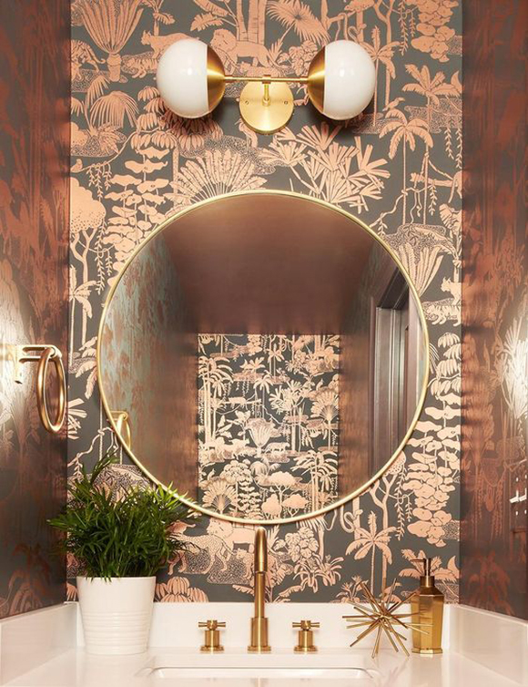 A 1930s-style powder room includes gold faucet and taps, a gold towel ring on the left wall, a large circular mirror on the rear wall, atop copper and black tropical jungle-themed wallpaper. Above the mirror is hung a double-pendant lamp with white globe shades. A green plant in a white vase adorns the countertop next to the taps.