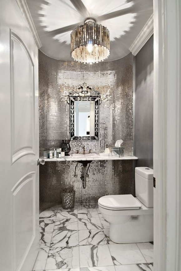 A beautiful curved powder room features white and grey marbled flooring, reflective mosaic wall tiling that bounces light from a round pendant light, and a glamorous silver mirror above a white marble sink and counter. A modern white toilet is visible in the front at the right, while the open white door appears at the left, framing the space.