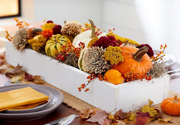 Photo of a white rectangular container in the center of a dining table, filled with small pumpkins, dried plants, pine cones, and scattered leaves on the table around it.