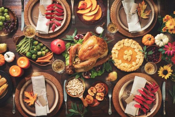 Overhead photo of a Thanksgiving meal spread out over a dark wood table, with turkey, pie, apple slices, artichokes, carrots, grapes, and plenty of candles and flowers spread throughout.
