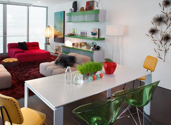 An open-plan living dining area as viewed from behind the white dining table with bright green acrylic chairs on silver metal legs, with 2 yellow head chairs. Across the room, a set of lime green shelves and storage units adorn the wall, while red and pink sofas sit atop a plush red rug.