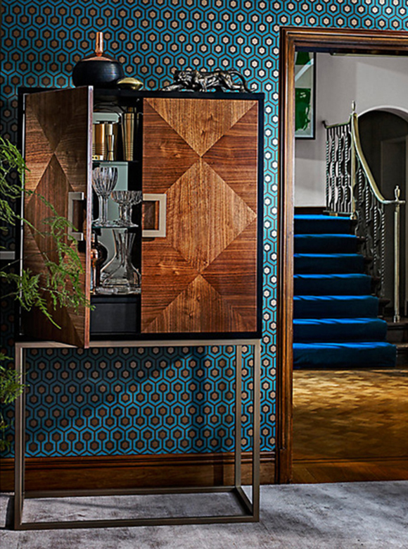 A peacock blue and gold hexagonal patterned wallpaper covers the walls behind a burled wood drinks cabinet with one open door revealing glasses and mixers inside.
