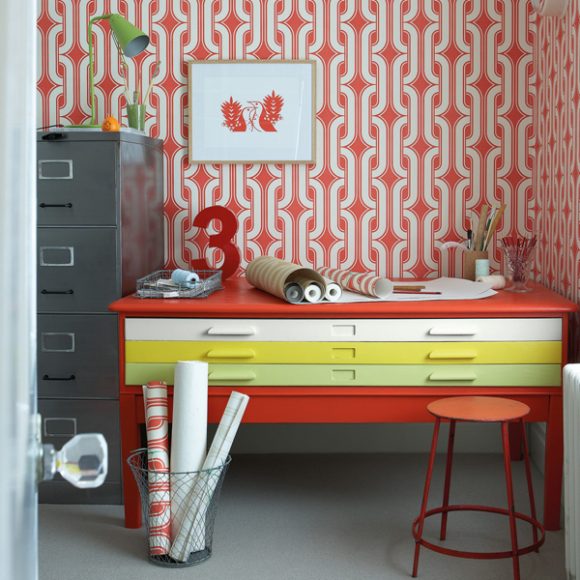 Photo of a corner of an office space, with a long red desk with 3 drawers painted in 3 colors of yellow, light green, and white. Patterned wallpaper in red and white covers the walls and sits in rolls on top of the desk. A red stool sits in front, with a metal bin also filled with wallpaper nearby.