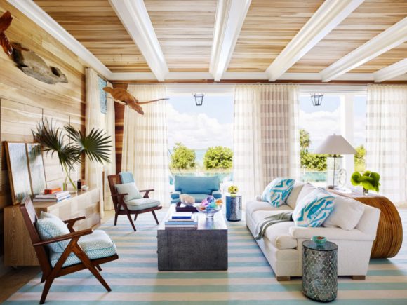 Looking into a living room with wood ceilings and walls, gauzy striped white and sand-colored curtains, light blue-green and white striped rug, a white sofa across from dark wood chairs with coordinating light-blue-green upholstery.