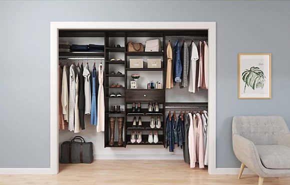 View into a neatly organized closet, with handing rails for clothes, racks for shoes, shelves for purses, and a drawer. Everything is spaced evenly, and easy to see. The walls outside the closet are grey, with a small framed image of a palm frond and a grey armchair to the right side.