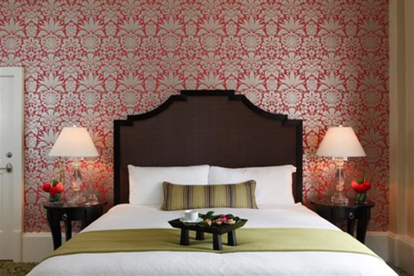 A dark wood headboard and bed dressed in white sheets with olive green striped pillow and matching green blanket across the foot of the bed, dressed with a dark wood serving tray. Behind the bed is a bold red and gold patterned wallpaper of pineapples. A pair of table lamps rest either side on dark wood bedside tables.