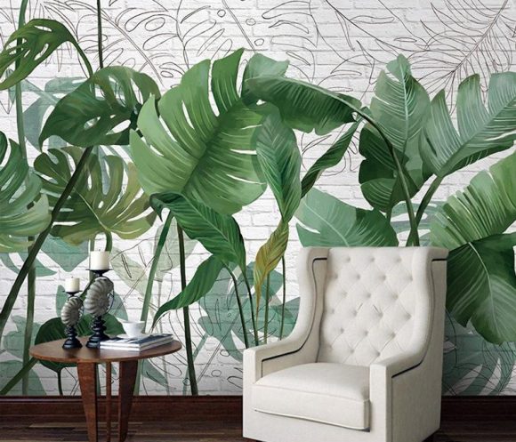 Photo of a realistic-looking wall mural of tropical fronds and plants, behind a white tufted upholstered armchair, next to a small wooden table dressed with books and a teacup.