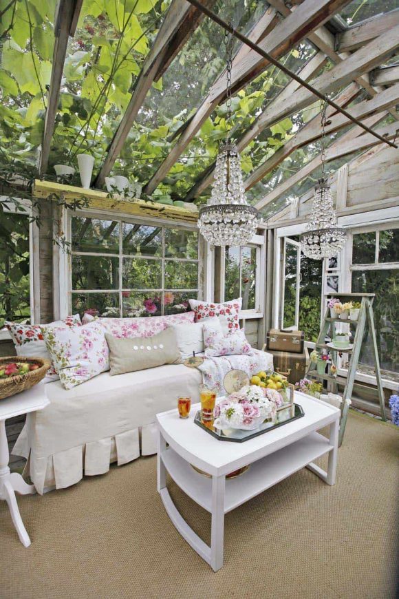 A glass-ceiling shed displays a tan slipcovered sofa with pink floral throw pillows and a white coffee table with a glass serving tray of lemonade sit underneath two fancy glass chandeliers. A ladder is used as shelves to display fresh flowers.