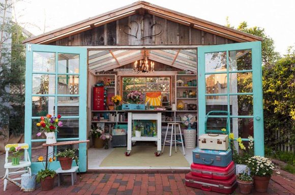 Two aqua-colored window pane doors hang open, revealing a table on wheels, a stool, multiple shelves with neatly organized crafting and painting supplies, all underneath a glass roof.
