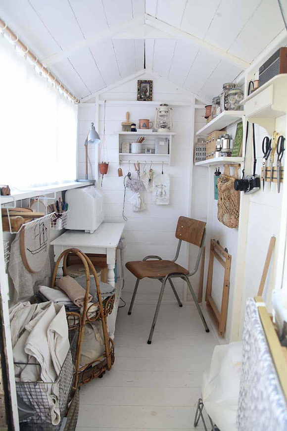 An all-white shed contains a sewing machine, chair, table, shelves with bottles of buttons, books and supplies, magnetic strips with scissors, and baskets of fabric are neatly organized under a large window with sheer curtains.