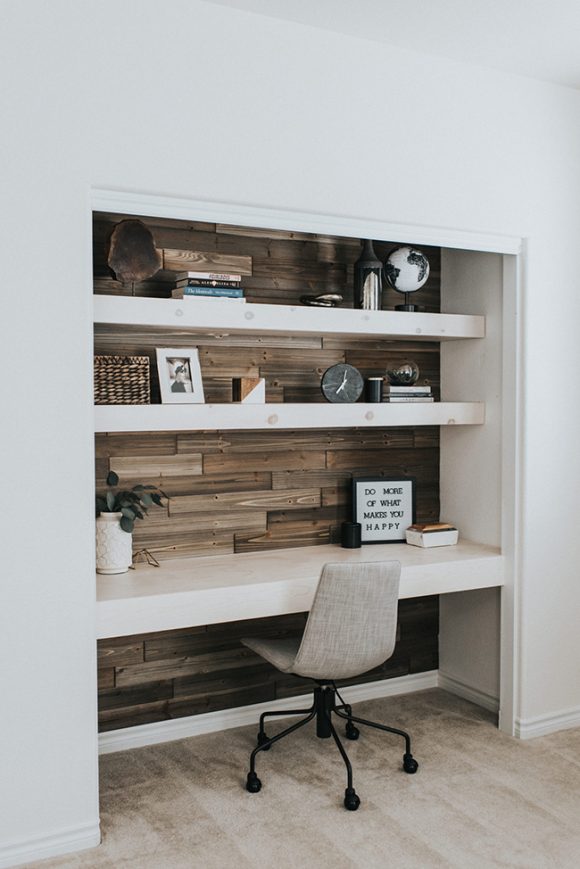 An elegant pocket office is created inside a bedroom closet by addition of a long chunky white wooden desk surface, two matching storage shelves above, and beautiful horizontal wood paneling on the back wall. A simple grey textured armless office chair on black casters completes the space. The shelves are dressed with photos, a clock, a globe, books, and textured baskets for office supplies.