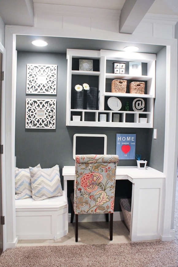 A wall nook turned into a home office area, with white built-in seating and pale herringbone pillows on the left, attached to a small white corner desk on the right with a pale blue, tan, red, and pink floral chair with dark wooden legs. Above the desk are multiple open shelves in white with cubbies and baskets to store office supplies. The rear wall is painted a dark chalky blue-grey, hung with white pierced wooden photo frames that match the desk and shelving.
