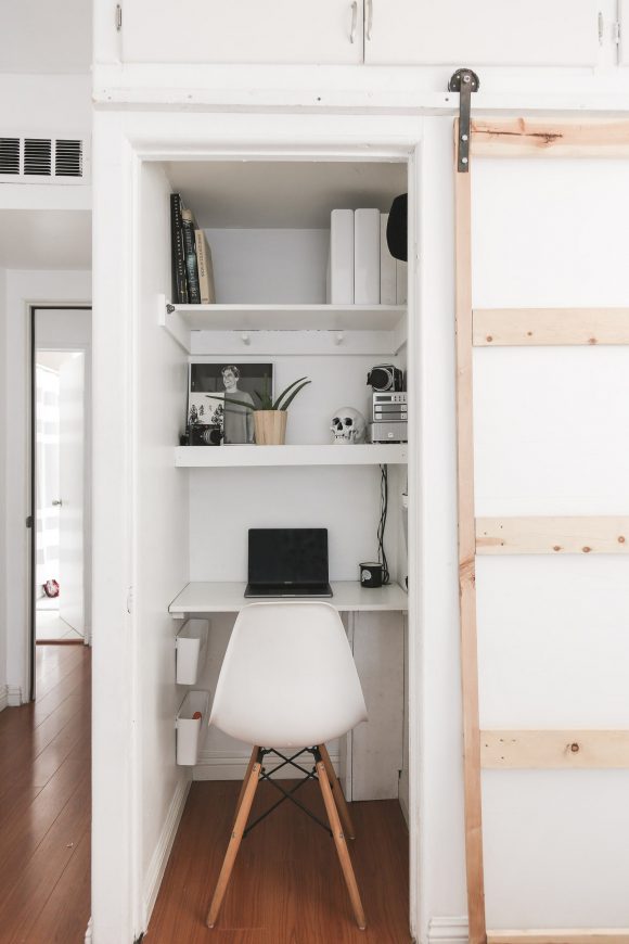 A linen closet is converted to a pocket office by adding deep shelving for storage and desk space, and a simple white molded fiberglass, metal and red wooden legged chair that tucks into the closet, whose door has been removed.