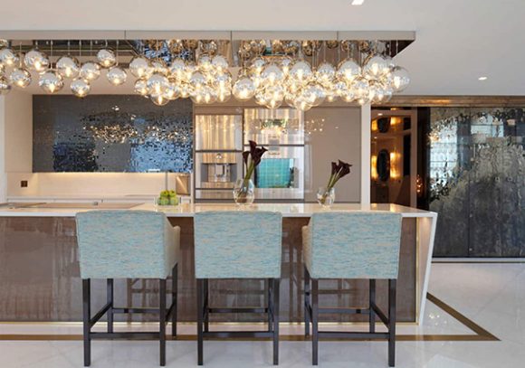 Image of a contemporary kitchen with sleek modern design, light colored countertop with a darker base and 3 blue and seafoam green colored chairs underneath a multiglobe chandelier that looks like bubbles.
