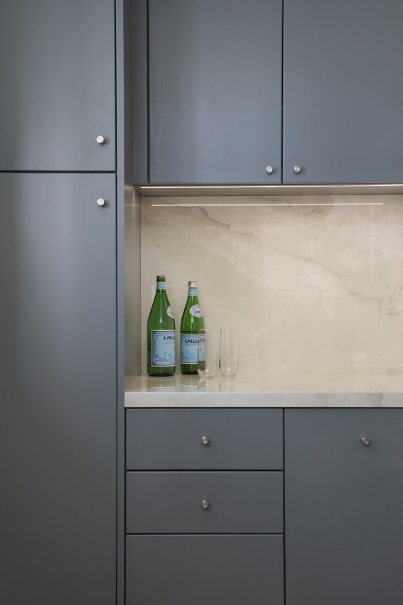 Sleek gray-green cabinetry with round silver knobs surrounds a white marble backsplash and countertop, dressed with 2 green bottles of Pellegrino water and clear glasses.