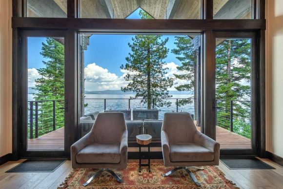 Two grey swivel armchairs sit in front of a floor-to-ceiling triple window with views of Lake Tahoe, trees and water. A deck and simple black railing expand the inside view outside.