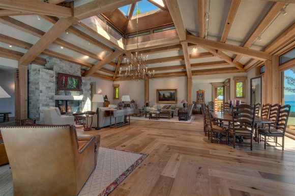 View of a large, open octagonal hardwood living room, with warm wooden ceiling joists creating a pattern above. 90-degree angled wood flooring complements the ceilings, while a large dining area, living area, and conversation area benefit from the views out the skylight.