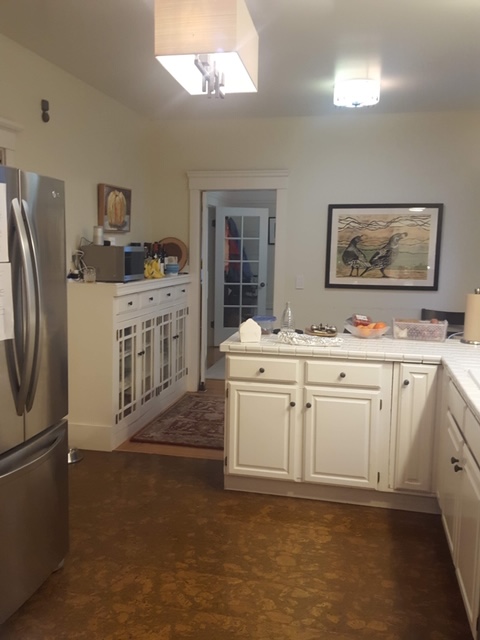 A dated, tired-looking kitchen with "country-style" cabinetry, worn flooring, and a large silver fridge/freezer combo. A small doorway at the end shows another room.