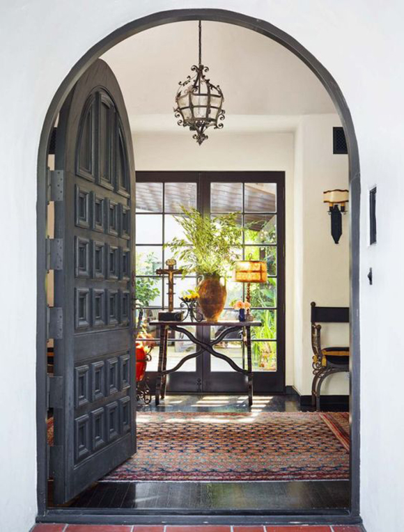 Coffered arched heavy wood front door opening onto a colorful rug, criss-crossed metal supports on a table and chair, in front of french doors showing greenery and streaming light, under a metal scrollwork pendant lamp in the entryway.