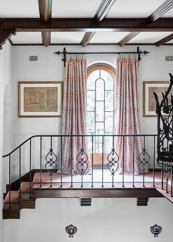 A staircase railing in delicate wrought iron scrollwork, in front of a pair of multicolored pink and purple and orange drapes, beneath dark wood beams, all in the Mission style.