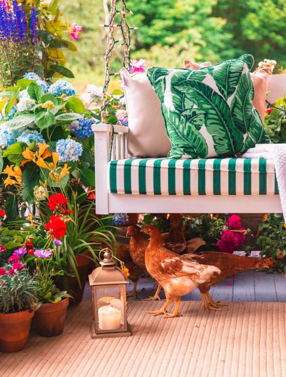 A white porch swing has green and white striped seat pillow, plus a green plant patterned throw pillow. Three chickens are underneath the swing, while colorful flowers and plants surround the porch. A copper portable lamp with candle sits on the porch floor, near the chickens.