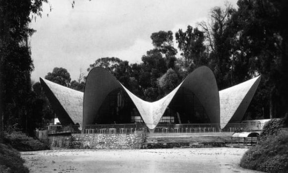 Black-and-white photo of the exterior of a large building with vaulted arch shapes that come down to touch the ground in between upward swoops, with chairs and tables inside. The outside is surrounded by trees.