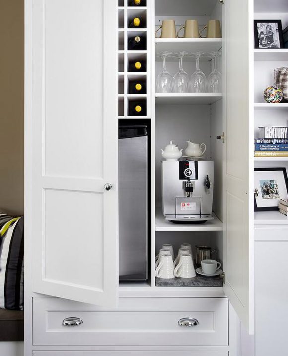 A pair of white cupboard doors open to reveal a small freestanding coffee maker, multiple cups and saucers, and on the top right shelf, wine glasses. A small wine fridge and bottle storage is just visible behind a partially open cupboard door. Underneath is a white drawer with silver handles.