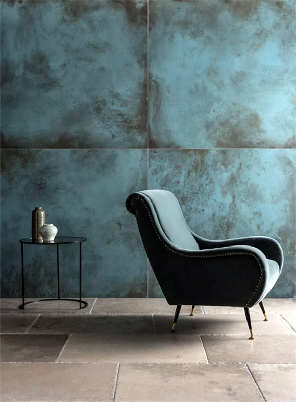 On the floor, oversized subway tiles in terracotta with white grout. On the walls, hugely oversized square tiles in turquoise blue with a rust patina. A matching turquoise armchair on 4 wooden legs faces away from a small black metal round side table dressed with two vases.