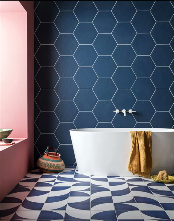 A colorful bathroom with blue and white wavy tiles on the floor, and large blue hexagons on the wall behind a white soaking tub with yellow towel hung over the side. To the right, the wall is rose pink all the way into the deep window seat.