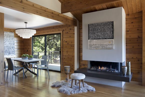 A warm knotty cedar wood living and dining area features a white custom-designed ribbon fireplace with a black hearth. A midcentury modern stool in white sits atop a fluffy grey faux fur in front of the fire. To the left, the dining area features a tiered white glass chandelier and a modern twisted white-topped pedestal table with grey-blue dining chairs. A large sliding glass door leads to the outside deck.