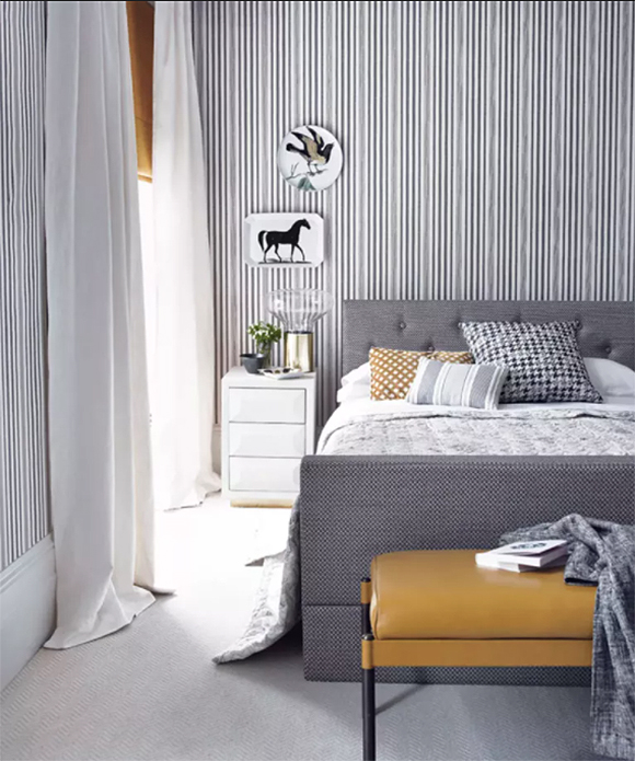 Black, white, and grey vertical stripes on wallpaper of this bedroom are softened by white draperies, punctuated with a mustard-yellow blind. A white side table sits next to a bed upholstered in tiny black-and-white checks. 3 pillows in varying sizes and shapes have black and white checks, grey stripes, and mustard-yellow patterning, respectively. A mustard-yellow footstool sits at the end of the bed, draped with a grey and white blanket and a couple of books.
