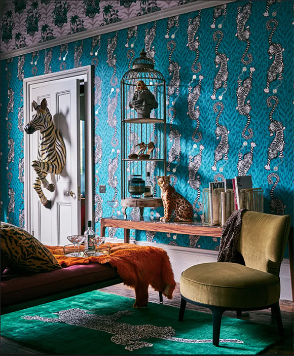 Bedroom with blue and black big-cat spotted wallpaper that also features white tigers crawling up and down. The front half of a zebra appears to leap from the back of the door, a birdcage hung on the wall holds a sculpture, pair of women's shoes, and perfume bottles on 3 levels used as shelves, sat upon a wooden console table with a wild cat sculpture next to magazine racks. Nearby, a hot pink chaise long is draped with an orange faux fur blanket and topped with a drinks server with cocktail glasses and a liquor bottle. On the floor, a bright green rug is layered with a black and white faux big cat skin, while a moss-green velvet chair nearby is draped with a black and white spotted shirt.