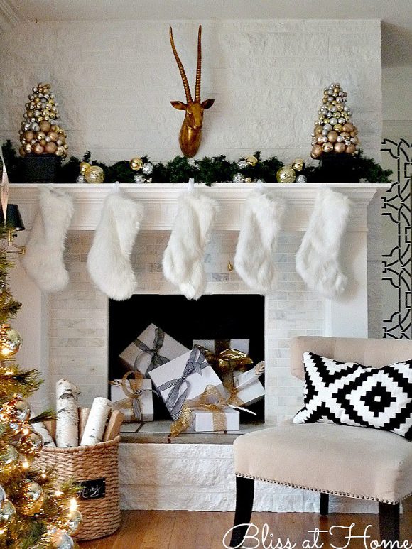 A white painted fireplace mantle and surround is covered in green pine swags plus gold and silver balls, and dressed with white furry stockings hung in a row. A pile of white gifts wrapped with silver and gold bows sits inside the fireplace, and a modern slipper chair in tan velvet with a black and white geometric pillow on the chair is off to the right, facing towards you. A hint of gold-covered Christmas tree is visible off to the left.