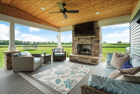 An outdoor living area with a stone-clad fireplace and TV above it, chairs and sofa with tan and light turquoise blue colors, matched in the center rug.