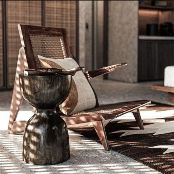 A low wooden chair with a white and rust pillow on it next to a dark brown drum-style side table, on a rug, on a concrete. In the background, concrete and woven material on vertical surfaces.