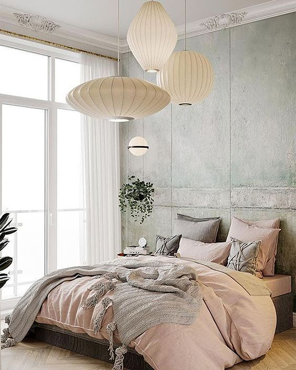 A bedroom featuring concrete walls is complemented by warm wooden flooring in a herringbone design. Pink and grey fabrics cover the queen bed with matching pillows. Above the bed hand 4 white paper lanterns in various shapes and sizes.