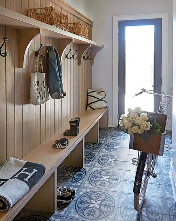 A wooden paneled wall with a long bench seat and spaces for tucking shoes away underneath sits atop a quatrefoil patterned tiled flooring in shades of blue. A bicycle whose basket is filled with flowers rests in front of the door, which has hazy light spilling in. Hooks on the wooden paneling for coats, hats, and scarves with a shelf above and baskets for capturing personal items.