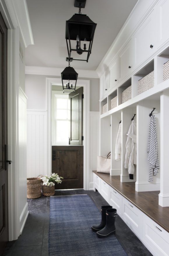 Wood paneling on the walls and trim all painted in white, while the ceiling and tops of the walls are in a beige grey. 5 cubbies have hooks for coats and a place for bags. Underneath is a drawer for shoes. Baskets at the top, with white cabinetry above. A wooden platform just above the shoe drawers allows people to sit to put their shoes on. A black stable door is open at the half, while a pair of black lamp light fixtures hang above. A blue rug runner makes the black tiling feel warmer.