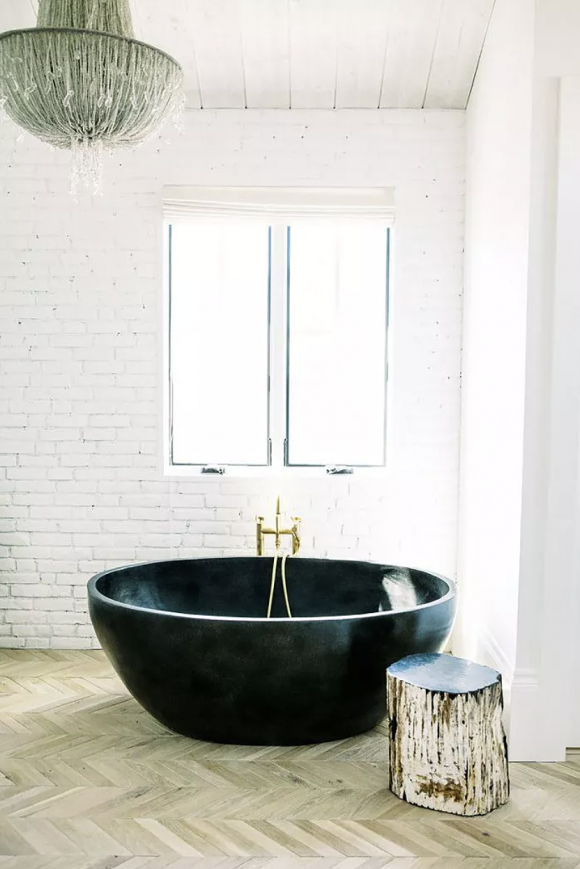 White painted subway tiles with a pair of plan windows set off the dramatic black soaking bathtub with brass fixtures, next to a real hewn tree as a side table, both sitting upon herringbone wood flooring in a variety of shades and colors, though tending towards the lighter end of the wood spectrum.